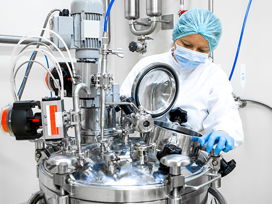 Alt-Text-Vorschlag: „Eine Person in Schutzkleidung, Haube, Mundschutz und Handschuhen arbeitet an einem großen Edelstahl-Reaktor in einem Labor oder einer Produktionsanlage.“