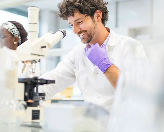 L&auml;chelnder Wissenschaftler arbeitet am Mikroskop, Kollegin im Hintergrund