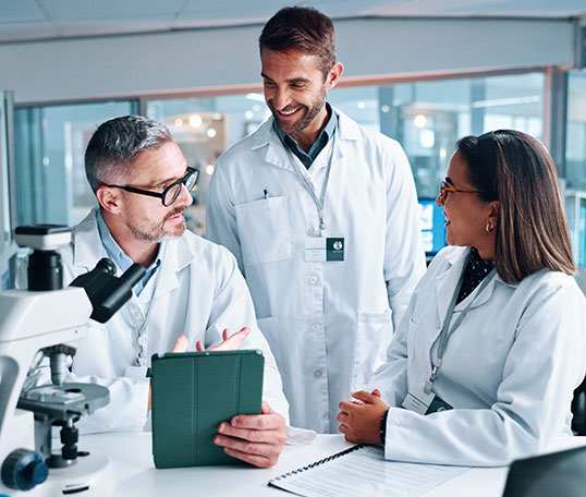 Drei Forschende in wei&szlig;en Laborkitteln stehen in einem modernen Labor zusammen, betrachten ein Tablet und besprechen Arbeitsergebnisse. Im Vordergrund ist ein Mikroskop zu sehen, im Hintergrund weitere Laborger&auml;te.