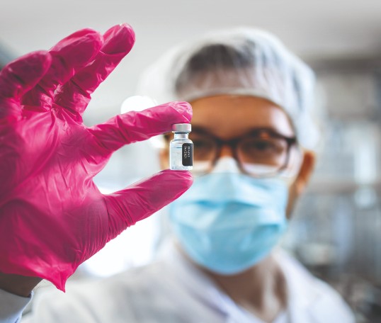 Drei Forschende in wei&szlig;en Laborkitteln stehen in einem modernen Labor zusammen, betrachten ein Tablet und besprechen Arbeitsergebnisse. Im Vordergrund ist ein Mikroskop zu sehen, im Hintergrund weitere Laborger&auml;te.