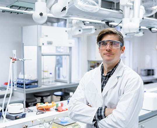 Ein Mitarbeiter in Laborkleidung und Schutzbrille steht in einem modernen Forschungslabor und blickt in die Kamera.